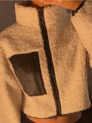  teddyjacka med svart detalj - Säljer en croppad teddyjacka i beige med svart dragkedja och en stor svart ficka på bröstet. Jackan har hög krage och är gjord i mjukt fleece-liknande material. Knappt använd 🩷