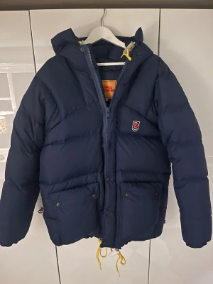 Fjällräven - EXPEDITION DOWN JACKET W - navy  - Säljer en mörkblå dunjacka från Fjällräven i storlek XXS. Jackan har huva, gula snören, stora fickor med knappar och klassisk Fjällräven-logga på bröstet. Perfekt för kalla dagar och riktigt skön att bära. Medföljer matchande påse. Mycket gott skick & varsamt använd ett fåtal gånger. Inga märken eller defekter. Ny pris 9 999kr.