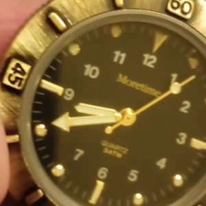 Moretime guldig klocka med läderband - Stilren klocka från Moretime med svart urtavla, guldiga visare och detaljer. Boetten är guldig och har markerade siffror på ringen. Klockan har ett brunt läderband som ger en klassisk look. Quartz-urverk och tydliga indexmarkeringar. Nytt batteri behövs.