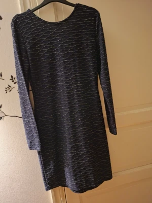 Mörkblå glittrig kortklänning - Säljer en mörkblå kortklänning med tunna glittriga vågmönster. Klänningen har lång ärm och v-ringning i ryggen, vilket ger en snygg och modern look. Perfekt för dig som vill sticka ut lite extra. Materialet känns mjukt och följsamt.