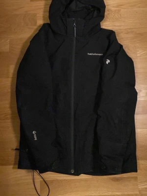 Svart vindjacka Peak Performance 160 - Svart vindjacka från Peak Performance med huva och hel dragkedja framtill. Jackan har diskret logga på bröstet och är tillverkad i slitstarkt syntetmaterial. Perfekt för blåsiga dagar och outdoor-aktiviteter. Snygg och enkel design som funkar till det mesta.