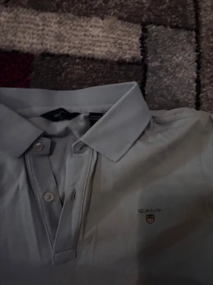 Ljusblå pikétröja från GANT - Snygg ljusblå pikétröja från GANT med klassisk krage och knappar framtill. Tröjan har korta ärmar och GANT-logga broderad på bröstet. Tillverkad i mjuk bomull som känns skön mot huden. Perfekt till jeans eller shorts.