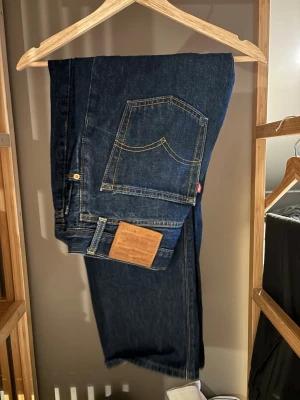 Levi's 501 mörkblå jeans - Klassiska Levi's 501 jeans i mörkblå tvätt med raka ben och gul kontrastsöm. Jeansen har fem fickor, knappgylf och den ikoniska läderpatchen bak i midjan. Tillverkade i slitstarkt bomullsjeansmaterial. Perfekta för en tidlös och avslappnad look.