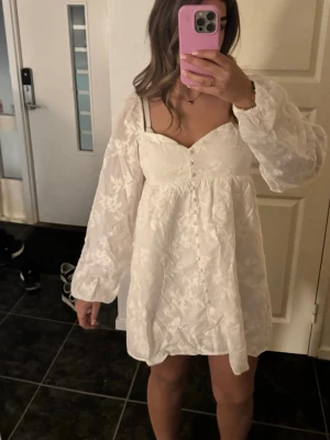 Vit kort klänning med blommönster - Supersöt vit kortklänning med blommigt mönster och v-ringning. Klänningen har långa puffärmar, knappar framtill och en luftig passform. Materialet känns lätt och flowy, perfekt för sommarens alla tillfällen.