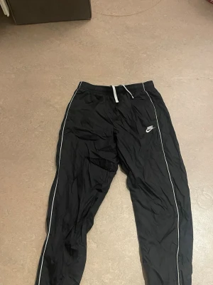 Svarta Nike täckbyxor med vita detaljer - Säljer ett par svarta täckbyxor från Nike med vita kontrastsömmar längs benen och vit dragsko i midjan. Byxorna har en sportig look och elastisk midja för skön passform. Perfekta för dig som gillar streetwear och vill ha något bekvämt och stilrent.