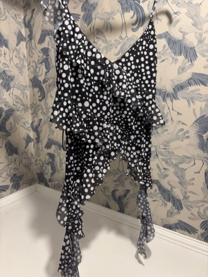 Svart prickig volangtop med asymmetrisk fåll - Säljer en svart topp med vita prickar och tunna axelband. Toppen har flera lager volanger och en asymmetrisk, fladdrig fåll som ger en cool och lekfull look. Materialet är lätt och meshigt, perfekt för dig som gillar statement-plagg. Super populär topp från Zara