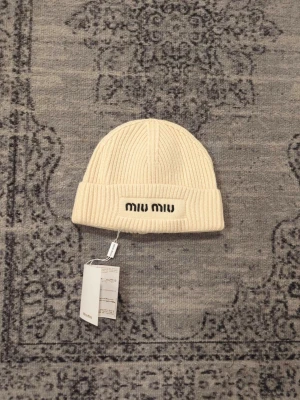 Beige ribbstickad mössa från Miu Miu - Säljer en beige ribbstickad mössa från Miu Miu med uppvikt kant och broderad logga framtill. Mössan är i mjuk ull och har en enkel, stilren design som passar perfekt till kyliga dagar.