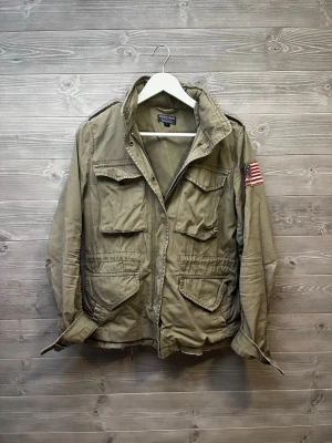 Olivgrön fieldjacka Polo Jeans Company - Säljer en olivgrön parkajacka från Polo Jeans Company, Ralph Lauren. Jackan har fyra stora fickor framtill, axelklaffar och en amerikansk flagga på ena ärmen. Tillverkad i bomull och har dragkedja framtill. Den är välanvänd med några skavanker men vilket ändast framhäver den nötta looken. 