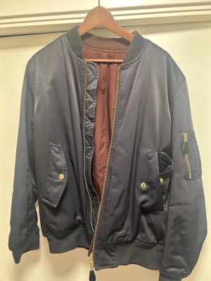 Svart bomberjacka från A.O.CMS - Säljer en svart bomberjacka från A.O.CMS med brunt innerfoder. Jackan har guldiga dragkedjor, fickor med tryckknappar och en klassisk ribbad krage och muddar. Perfekt för dig som vill ha en stilren och enkel jacka med snygga detaljer. Jackan är vändbar.