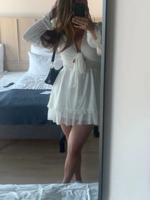 Vit kort omlottklänning med knyt - Söt vit kortklänning med omlottdesign och knyt framtill. Klänningen har långa ärmar i ett lätt, flödande material och volangdetaljer längst ner. Perfekt för sommarens alla tillfällen och ger en romantisk vibe.