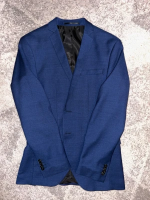 Blå kavaj set med kostymbyxor från Jack & Jones - Snygg marinblå kavaj från Jack & Jones i super slim passform. Kavajen har klassisk krage, tre fickor fram och fyra svarta knappar vid ärmslutet. Materialet är troligtvis polyester och kostymen  har en stilren look som passar vid lite finare tillställningar. Hör av er vid intresse/ frågor!