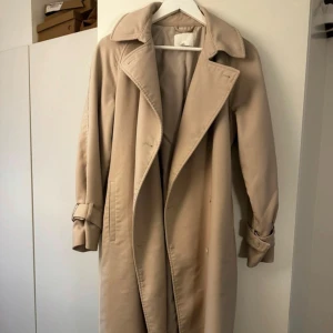 Beige trenchcoat med bälte - Säljer en klassisk beige trenchcoat med bred krage, knytskärp i midjan och detaljerade ärmslut med spännen. Jackan har en rak passform och är tillverkad i ett mjukt tyg som känns skönt att bära. Perfekt för dig som vill ha en stilren och tidlös look. Den har även tjockare material än en vanlig trenchcoat!