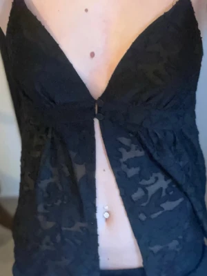 Svart spets topp med knappar - Säljer en svart topp i transparent spets med djup v-ringning och smala axelband. Toppen har två knappar framtill och ett lätt utställd passform. Perfekt för dig som vill ha en edgy och trendig look.