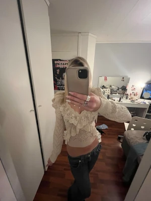 Beige stickad blus med volang - Supersöt beige stickad blus med volangdetaljer och v-ringning. Blusen har långärmade ärmar och är croppad, med spets och broderade detaljer som ger en romantisk vibe. Perfekt att styla med jeans eller kjol för en trendig look.