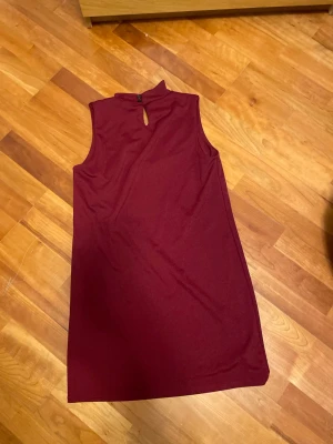 Vinröd ärmlös klänning från Boohoo - Säljer en vinröd, ärmlös kortklänning från Boohoo i storlek 40. Klänningen har en liten ståkrage med knappdetalj och en snygg öppning bak i nacken. Materialet känns mjukt och stretchigt, perfekt för dig som gillar enkel men stilren design.