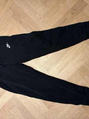 Svarta mjukisbyxor från Nike, stl S - Svarta mjukisbyxor från Nike med klassisk logga på vänster lår. Byxorna har resår i midjan och vid bensluten samt dragsko för justerbar passform. Tillverkade i mjukt material som är skönt att ha på sig. Perfekta för chill eller träning.