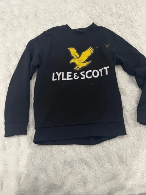 Svart sweatshirt från Lyle & Scott - Svart sweatshirt från Lyle & Scott med stor gul logga och text på bröstet. Klassisk rund halsringning och långa ärmar. Tillverkad i mjuk bomullsmix, perfekt för chill dagar. Ribbstickade muddar vid ärmslut och nederkant. Ändå felet men den är att den har klorin fläckar på sig 
