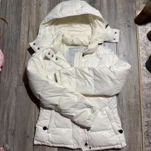 Vit pufferjacka från Benetton - Säljer en vit pufferjacka från Benetton med huva och dragkedja. Jackan är vadderad med 80% dun och 20% fjäder, har tryckknappar och två fickor framtill. Perfekt för kalla dagar och har en clean, modern look. ‼️ OBS att en del av hänget som man drar i vid dragkedjan är borta men det hindrar inte funktionen