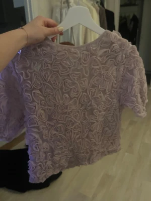 Ljuslila volangblus med rosdetaljer - Supersöt ljuslila blus med korta ärmar och massor av volangiga rosdetaljer över hela tyget. Blusen har en boxig passform och är perfekt för dig som vill sticka ut med en unik och romantisk look.