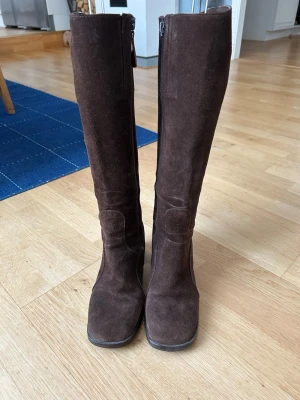 Bruna mockastövlar med klack - Snygga bruna stövlar i mocka från Aneto med fyrkantig tå och bred klack. Dragkedja på insidan av skaftet gör dem enkla att ta på. Perfekta till höst- och vinterlooken, matcha med jeans eller kjol för en trendig vibe.