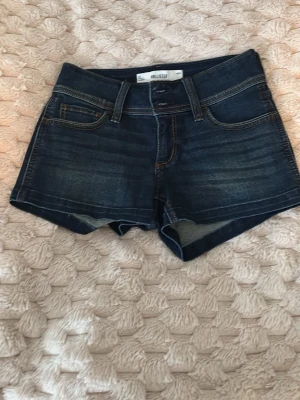 Mörkblå low-rise jeansshorts Hollister - Säljer ett par mörkblå low-rise jeansshorts från Hollister. Klassisk femficksmodell med snygga bakfickor med lock och knapp. Shortsen har kontrastsömmar och är perfekta för sommaren. Passar dig som gillar en avslappnad och trendig stil.