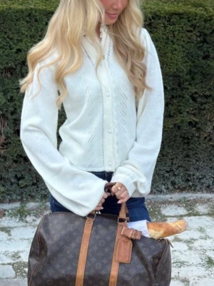 Meet me there x Filippa Montan tröja - Superfin vit stickad kofta med dekorativa spetsdetaljer längs knäppningen och halsen. Koftan har långärmade puffärmar, fina mönsterstickningar och pärlknappar framtill. Perfekt för lager-på-lager och ger en romantisk vibe till din outfit.