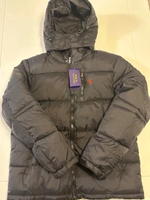 Svart pufferjacka från Polo Ralph Lauren - Säljer en svart pufferjacka från Polo Ralph Lauren med huva och dragkedja framtill. Jackan har det klassiska röda Polo-logot på bröstet och är tillverkad i ett glansigt syntetmaterial. Perfekt för kalla dagar och riktigt snygg till streetwear-stilen.