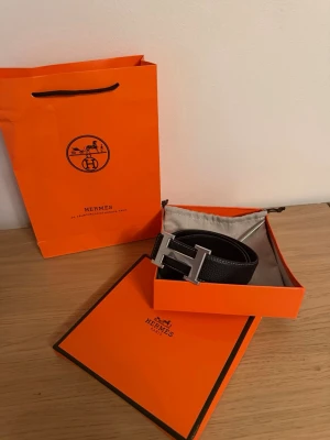 Svart bälte - Säljer ett svart bälte med det ikoniska H spännet i silverfärg. Bältet är gjort i exklusivt skinn med vit kontrastsöm och levereras i originalförpackning med dustbag och påse. Perfekt accessoar för att lyfta vilken outfit som helst.