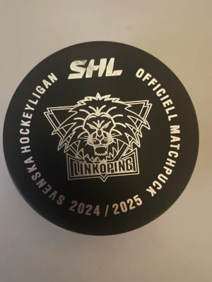 SHL Linköping officiell matchpuck - Säljer en svart officiell matchpuck från SHL säsongen 2024/2025 med Linköpings HC:s lejonlogo i vitt tryck. Pucken har tydlig SHL-logga på ena sidan och text runt kanten. Perfekt för dig som samlar på hockeyprylar eller vill ha ett coolt minne från säsongen.