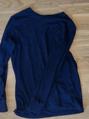 Mörkblå långärmad tröja Polo Ralph Lauren - Mörkblå långärmad tröja från Polo Ralph Lauren med klassisk röd broderad logga på bröstet. Tillverkad i mjuk bomull som känns skön mot huden. Perfekt till chill dagar eller när du vill ha en clean och enkel look.