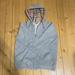 Grå hoodie från Burberry med dragkedja - Säljer en grå hoodie från Burberry med dragkedja och klassiskt rutigt foder i huvan. Tröjan har fickor framtill och broderad logga på bröstet. Perfekt för en avslappnad och stilren look.
