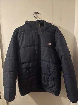 Mörkblå pufferjacka från Levi's - Säljer en mörkblå pufferjacka från Levi's i storlek S. Jackan har huva, två sidofickor och dragkedja framtill. Klassisk quiltad look och liten Levi's-logga på bröstet. Perfekt för kyliga dagar och enkel att matcha med olika outfits.