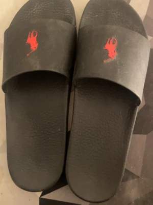 Ralph Lauren tofflor  - Svarta tofflor från Polo Ralph Lauren med röd logga framtill och präglad sula för extra grepp. Klassisk design med bred rem över foten och Polo-tryck undertill. Tillverkade i syntetmaterial, perfekta för sommardagar eller chill hemma. 