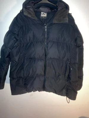  Svart McKinley pufferjacka - Säljer en svart pufferjacka från McKinley i storlek 46. Jackan har huva, dragkedja framtill och två sidofickor, och två innerfickor. Perfekt för kalla dagar med sin vadderade design och klassiska look. Passar dig som vill ha en varm och enkel jacka till vintern. Jackan har dock två skador som är lagade (kolla bilder)