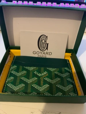 Grön Goy4ard plånbok med mönster - Säljer en grön plånbok från Goy4ard med deras ikoniska grafiska mönster i vitt och beige. Plånboken har flera kortfack och är tillverkad i skinn. Kommer i originalask med Goy4ard-logga och gul insida. Perfekt för dig som vill ha något riktigt stilrent och klassiskt.