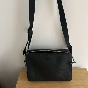 Svart axelväska från Lacoste - Snygg svart axelväska från Lacoste med diskret krokodillogga på framsidan. Väskan har justerbar rem, silverfärgade dragkedjor och ett strukturerat yttertyg. Insidan är fodrad i grönt tyg med innerfack och dragkedja. Perfekt för att bära det viktigaste.