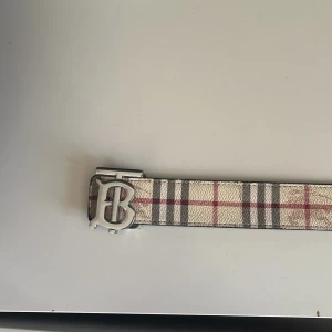 Burberry bälte med klassiskt rutmönster - Säljer ett snyggt Burberry bälte med ikoniskt rutmönster i beige, svart och rött samt en stor silvrig B-spänne. Bältet är gjort i skinn och har en exklusiv känsla. Perfekt accessoar för att lyfta din outfit med en touch av lyx.