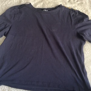 Mörkblå topp med guldknappar H&M - Mörkblå topp från H&M Divided med rund halsringning och tre snygga guldknappar på ena axeln. Tillverkad i mjuk bomull och har korta ärmar. Perfekt för dig som gillar stilrena detaljer och enkel design.