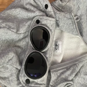 Grå hoodiejacka från C.P. Company - Säljer en grå hoodiejacka från C.P. Company med ikoniska goggles i huvan. Jackan har dragkedja framtill, snörning i huvan och är tillverkad i mjuk bomull. Perfekt för dig som vill sticka ut med streetwear-vibe och coola detaljer.
