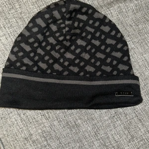 Svart och grå mönstrad mössa från BOSS - Snygg svart och grå mössa från BOSS med coolt geometriskt mönster och diskret logga framtill. Tillverkad i Italien och stickad i mjukt material som håller dig varm. Perfekt accessoar för att lyfta din stil under kyliga dagar.