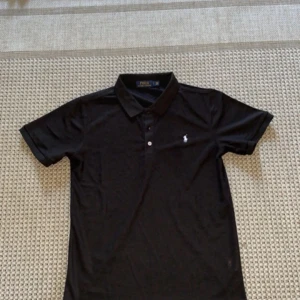 Svart pikétröja från Polo Ralph Lauren - Klassisk svart pikétröja från Polo Ralph Lauren med vit broderad logga på bröstet. Tröjan har krage, knappar framtill och korta ärmar. Perfekt för en clean och stilren look.