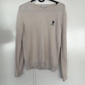 Beige stickad tröja U.S. Polo Assn S - Snygg beige stickad tröja från U.S. Polo Assn. med diskret logga broderad på bröstet. Tröjan har långärmad passform, rund halsringning och ribbade muddar. Perfekt för lager-på-lager och enkel att matcha med jeans eller kjol.