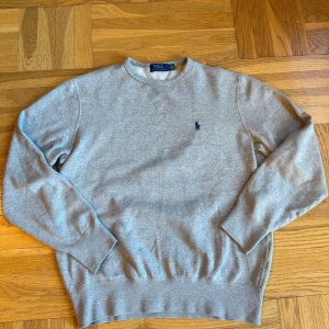 Grå sweatshirt från Polo Ralph Lauren - Klassisk grå sweatshirt från Polo Ralph Lauren med rund halsringning och broderad marinblå logga på bröstet. Tröjan har ribbade muddar vid ärmslut och nederkant, perfekt för en clean och avslappnad stil. Tillverkad i mjuk bomullsmix.