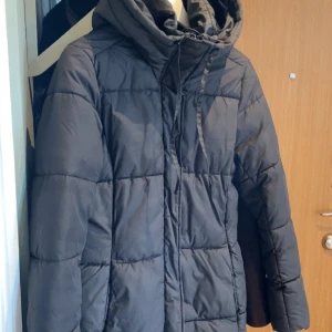 Svart pufferjacka med huva XS - Säljer en svart pufferjacka i storlek XS med stor, mysig huva och snörning. Jackan har quiltad design och långa ärmar, perfekt för kalla dagar. Är mer svart i verkligheten. Lång jacka
