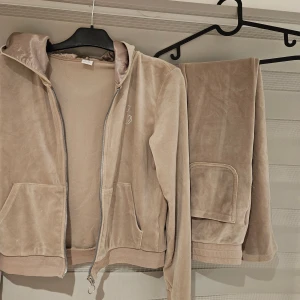 Beige velour hoodie  med byxa från Lindex - Mjuk och skön beige hoodie i velour med byxa från Lindex med glansig huva och dragkedja. Framsidan har en snygg strassdetalj i form av ett C. Perfekt för dig som gillar en chill och trendig stil.