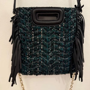 Axelväska från Maje med fransar - Snygg axelväska från Maje i mörkgrönt och svart tweedtyg med coola fransdetaljer i skinn på sidorna. Väskan har ett rektangulärt handtag upptill i svart och kedjerem. Perfekt för dig som vill sticka ut med en unik accessoar.