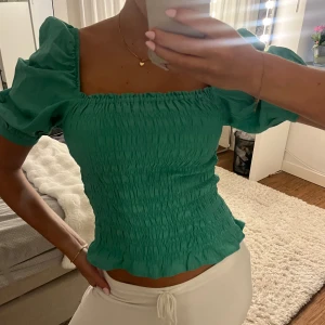 Grön smockad blus med puffärm - Supersöt grön blus med smockad kropp och puffiga korta ärmar. Fyrkantig urringning och lätt volangkant nedtill ger en trendig och feminin vibe. Perfekt att styla med jeans eller kjol för en fräsch look.