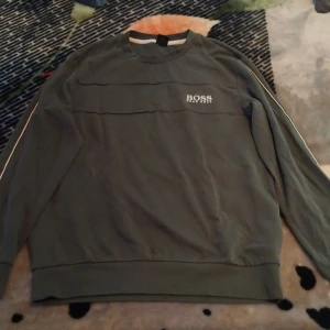 Gröna mjukisbyxor och tjock tröja från Hugo Boss M - Snygga gröna trachsuit från Hugo Boss i storlek M. Byxorna har vita ränder längs sidorna samma som på tröjan, snörning i midjan och ribbade muddar vid bensluten. Boss-logga tryckt på ena benet. Perfekta för chill eller streetwear.