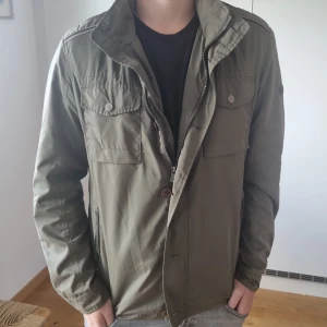 Säljer denna feta olivgröna vindjackan, hör av er vid minsta fundering  - Snygg olivgrön vindjacka från Devota & Lomba med dragkedja och knappar framtill. Jackan har två bröstfickor med lock och knappar samt två sidofickor med dragkedja. Fodret är randigt i blått och vitt, vilket ger en fräsch touch.
