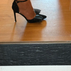 Svarta pumps med rosett och klack - Säljer ett par svarta pumps med spetsig tå och hög klack. Skorna har en snygg rosettdetalj framtill i mesh och smal ankelrem med spänne. Materialet är mockaliknande och de är perfekta för dig som vill sticka ut lite extra.
